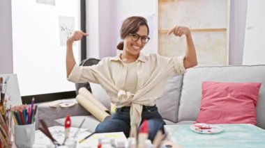 Kendinden emin, çekici bir sanatçı olan genç İspanyol kadın stüdyo kanepesinde gururla kendini işaret ediyor, mutlu görünüyor.