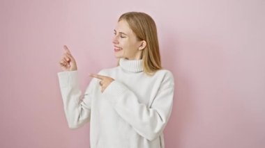 Neşeli, güzel sarışın kız kendinden emin bir şekilde yan tarafı işaret ediyor, izole edilmiş pembe bir duvarın karşısında duruyor, kameraya gülümsüyor.