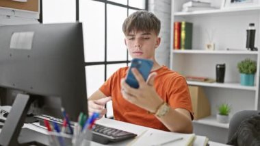 Turuncu tişörtlü beyaz erkek öğrenci modern bir üniversite kütüphanesinde akıllı telefonuyla selfie çekiyor..