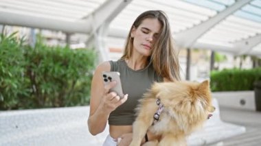 Genç, kendine güvenen İspanyol kadın neşeyle gülümsüyor, güzel köpeğine sarılıyor, parkta telefonuyla neşeli bir şekilde mesajlaşıyor..