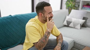 Sakallı ve dövmeli düşünceli İspanyol adam oturma odasında kanepede oturuyor.