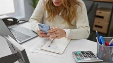 Modern ofis ortamında bilgisayar ve kırtasiyeyle akıllı telefon kullanan gülümseyen kadın..