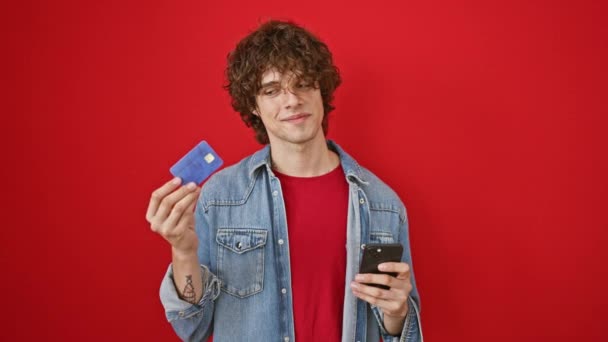 头发卷曲的男人拿着信用卡和红色背景的智能手机微笑 — 图库视频影像