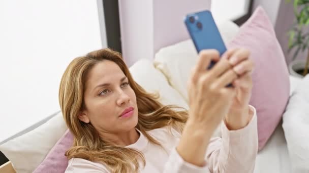 一个穿着休闲装的年轻女人在舒适 现代的卧室里拿着智能手机自拍 — 图库视频影像