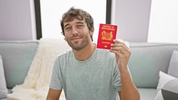 自信のある金髪の男は 自宅のソファーで微笑み 愛国的に休暇の準備ができている彼のシンガポールのパスポートを保持しています — ストック動画