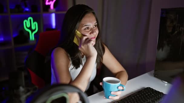 魅力的な若いヒスパニック系女性ストリーマーがゲームストリームに浸り スマートフォンで会話をしたり ネオンリットのゲームルームでコーヒーを飲んだりしました — ストック動画