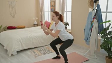 Güzel dekore edilmiş, rahat bir yatak odasında telefonuna günlük kıyafet tomarları takmış odaklanmış beyaz bir kadın..