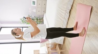 Aktif kıyafetli genç bir kadın, yoga minderiyle parlak bir yatak odasında akıllı bir telefonla takılıyor.