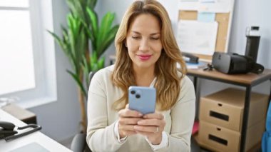 Gülümseyen bir kadın ofis masasında akıllı telefon kullanır. Bağlılık ve profesyonelliği ifade eder..