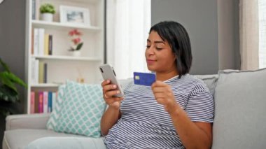 İspanyol kadın oturma odasındaki kanepede dinlenirken online alışveriş için akıllı telefon ve kredi kartı kullanıyor.