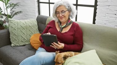 Gri saçlı olgun bir kadın rahat bir şekilde kanepede evcil köpeğiyle birlikte tablet kullanıyor..
