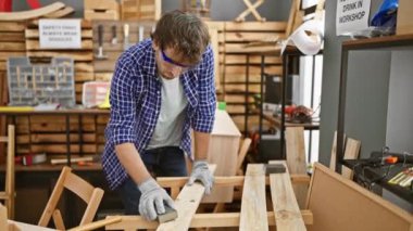 Yakışıklı, genç, sakallı, gözlüklü, mesleğine dalmış, ahşap bir kalas zımparalıyor marangozluk atölyesinde.