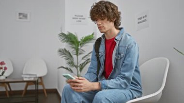 Dalgın, kıvırcık saçlı, kot ceket giyen genç bir adam, akıllı telefonuna bakarken beyaz bir bekleme odasında oturuyor..