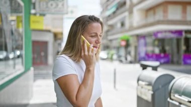 Şık genç sarışın kadın ciddi telefon konuşmalarına dalmış, güneşli şehir caddesinde dikiliyor, konsantrasyonunu gösteriyor, gelişigüzel, kentsel bir yaşam tarzını kusursuz bir şekilde yakalıyor..