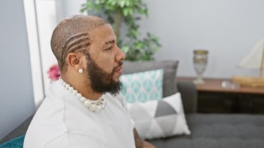 Afro-Amerikalı özgüvenli bir adam, doğal olarak gülümsüyor, evdeki bir koltukta yan oturuyor, yakışıklı bir profili ortaya çıkarıyor.