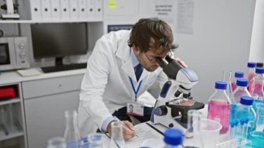 Ciddi bir genç bilim adamı, sakallı bir sarışın, laboratuvar masasında titizlikle notlar alıyor, tıp araştırmalarına mikroskop altında dalmış,.