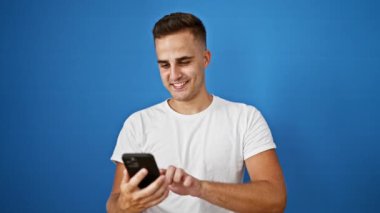 Gülümseyen genç İspanyol adam mavi duvara karşı akıllı telefon kullanıyor.