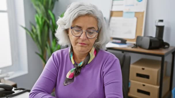成熟した女性は 明るいオフィスの設定で時間をチェックし メガネと紫色のセーターを着用します — ストック動画