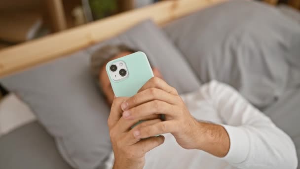 惊慌失措的男人 灰白的头发 舒服地躺在床上 专心致志地用智能手机发短信 他舒适的卧室里充满了清爽的早晨气氛 — 图库视频影像