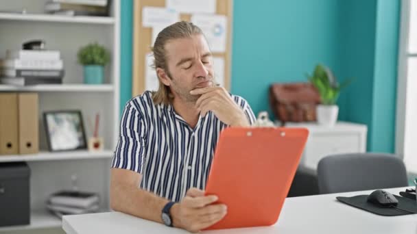 長髪の熟練した男性がモダンなオフィス内装のクリップボードを保持し 思慮深さを表現 — ストック動画