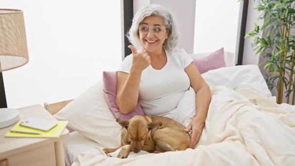 パジャマの陽気な中年の女性は 居心地の良い寝室で彼女の灰色の髪の犬とベッドに座っている間 親指を指して 広く笑います — ストック動画