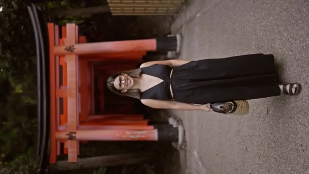 きれいな笑顔と眼鏡で 気楽で楽しい若いヒスパニック女性が 京都のオレンジ色の鳥居の門のカメラに向かって歩いていると自信を持ってポーズします — ストック動画