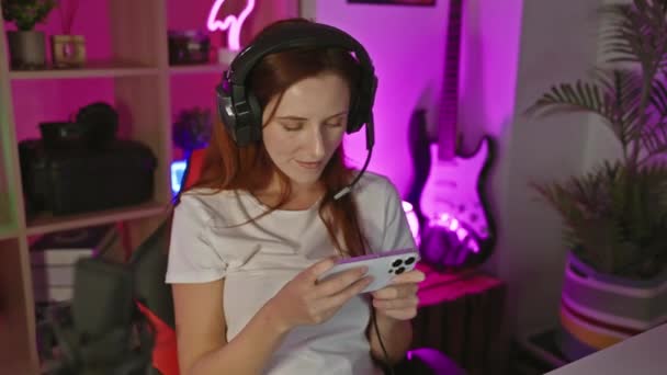 夜のネオンライトゲームルームでスマートフォンを使用してヘッドフォンを持つ笑顔の若い女性 — ストック動画