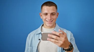 Yakışıklı genç adam mavi izole arka plana karşı akıllı telefon kullanıyor. Günlük kıyafetleri ve saati var..