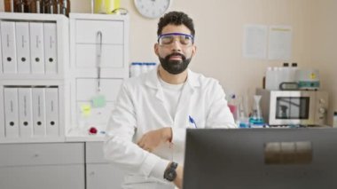 Laboratuvar önlüklü, sakallı yakışıklı genç adam tıbbi laboratuvar ortamında güvenle gülümsüyor..