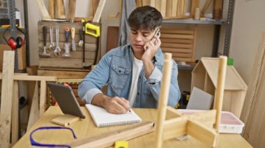 Genç, yakışıklı İspanyol marangoz atölyesinde çalışıyor, akıllı telefonuyla konuşuyor, kereste ve aletlerin arasında dokunmatik çizimler yapıyor.