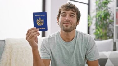 Işıldayan, kendine güvenen genç sarışın adam, oturma odasındaki kanepesinde oturmuş, Küba pasaportunu sallıyor, mutluluk ve neşe saçıyor, hayatının yolculuğuna hazır.