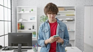 Şık, kıvırcık saçlı ve gözlüklü genç bir adam modern ofis ortamında akıllı bir telefondan mesaj atıyor..