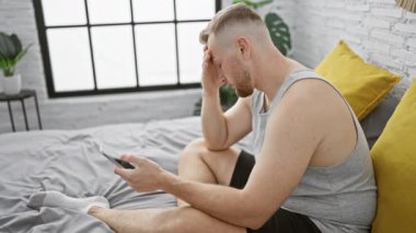 Askılı tişörtlü genç sakallı bir adam yatak odasında akıllı telefon tutarken yorgun görünüyor.