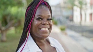 Neşeli Afro-Amerikan kadını, kendine güvenen ve gülümseyen, güzel yeşil bir şehir parkında sıradan duran, mutluluk saçan ve dışarıda eğlenirken pozitif titreşimler saçan