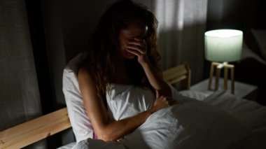 Sıkıntılı genç bir kadın loş bir odada yatakta oturuyor, yüzünü örtüyor ve ağlıyor..