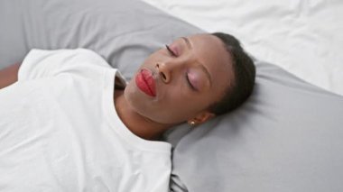 Şok olmuş Afro-Amerikalı bir kadın gözlerini ve ağzını şaşkınlıkla kapatıyor, yatakta uzanıyor, duygularını ustalıkla saklıyor.