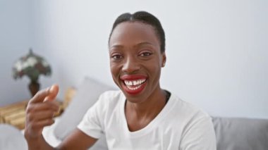 Gülümseyen Afro-Amerikalı kadın, parmaklarını yüzüne ve burnuna doğrultarak, yatak odasında uzanırken güzel bir ışık saçıyor.