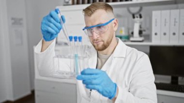 Laboratuvar gözlüklü ve eldivenli bir adam laboratuarın içinde test tüplerini inceliyor..