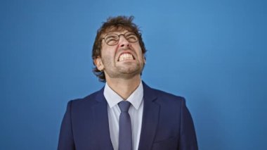 Öfke ve saldırganlıkla kaynayan takım elbiseli sinirli genç adam öfkesini haykırıyor. Mavi bir arka planda izole edilmiş, öfkenin titiz bir portresi..