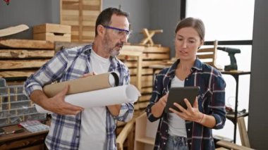 İki gülümseyen marangoz, erkek ve kadın, ahşap işi ortağı, dokunmatik yastık kullanıyor ve planları tutuyor, modern marangozluk ve inşaat endüstrisine bir göz atıyor.
