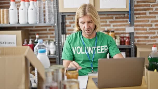 緑色のボランティアのシャツを着た長髪の男性が 寄付の棚のある倉庫でラップトップに取り組んでいる — ストック動画