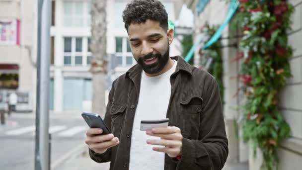 在繁忙的城市街道上 一个留着胡子 面带微笑的年轻人一边拿着信用卡一边看手机 — 图库视频影像