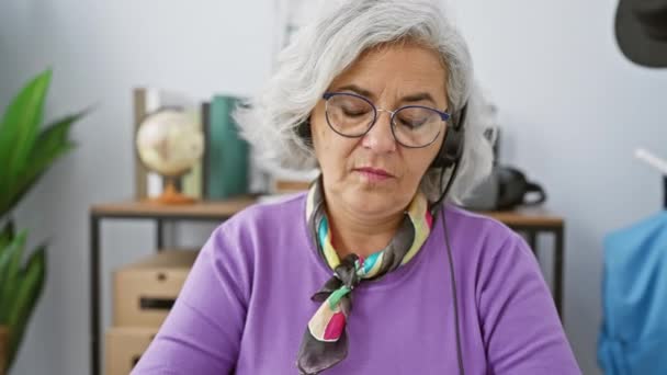現代のオフィスの設定でヘッドセットを着用した灰色の髪とメガネの笑顔の成熟した女性 — ストック動画