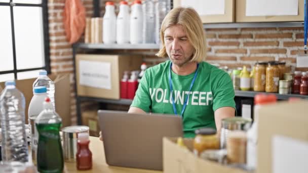 緑色の ボランティア Tシャツを着たブロンドの髪を持つ中年の男性は 食品銀行の倉庫でラップトップに注意深く取り組んでいます — ストック動画