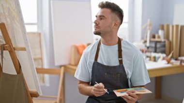 Önlük takan sakallı düşünceli genç bir adam bir sanat stüdyosunda boya paleti tutuyor..