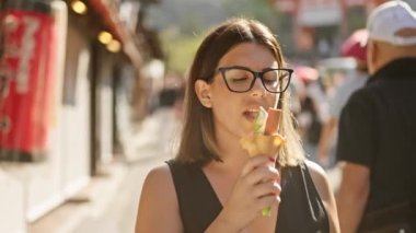 Neşeli İspanyol kadın Kyoto caddesinde nefis bir şekilde tatlı dondurmanın tadını çıkarıyor, güzel gülüşü bu Japon şehrinin geleneksel mimarisini deneyimlerken gözlüklerle çerçeveleniyor.
