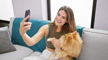 Rahatlamış genç İspanyol kadın cep telefonunda komik bir video görüşmesi yapıyor yanında mutlu köpeği, evdeki oturma odasındaki kanepede oturuyor..