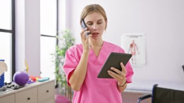 Pembe önlüklü sarışın kadın telefonda konuşuyor ve fizyoterapi kliniğinde tablet okuyor..