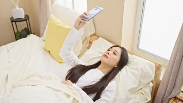 Überraschte Asiatin Liegt Mit Smartphone Bett Hellen Schlafzimmer — Stockvideo