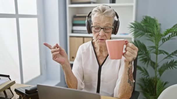 ハッピー 自信を持って上級女性労働者 灰色の髪 ヘッドフォン上の 音楽で失われた コーヒーを飲む 忙しいオフィスで彼女のラップトップで笑顔 — ストック動画
